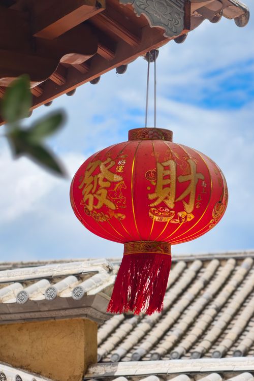 Free A vibrant red Chinese lantern with calligraphy hangs outdoors from a traditional roof. Stock Photo