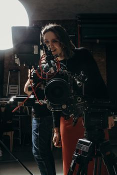 A female videographer adjusts studio camera equipment under soft lighting.