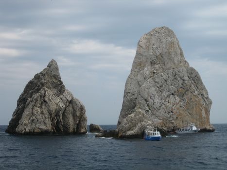 Two striking rock formations rise from the sea, framed by a cloudy sky.