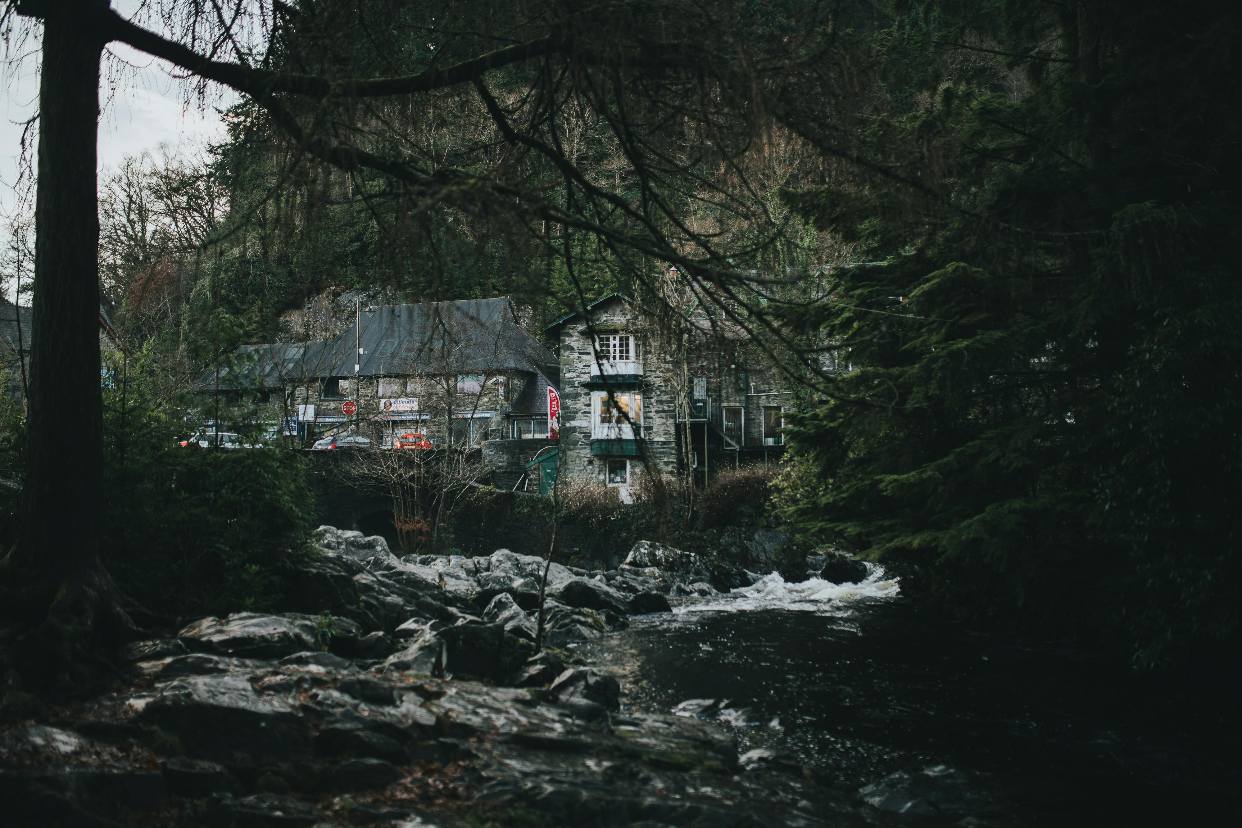 Rustic stone house nestled in lush forest alongside a flowing river in Wales, UK.