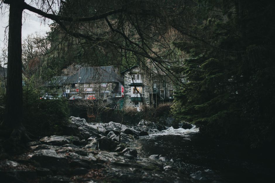 Rustic stone house nestled in lush forest alongside a flowing river in Wales, UK.