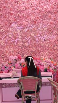 A woman sits in a floral cafe interior with pink decor and neon sign.