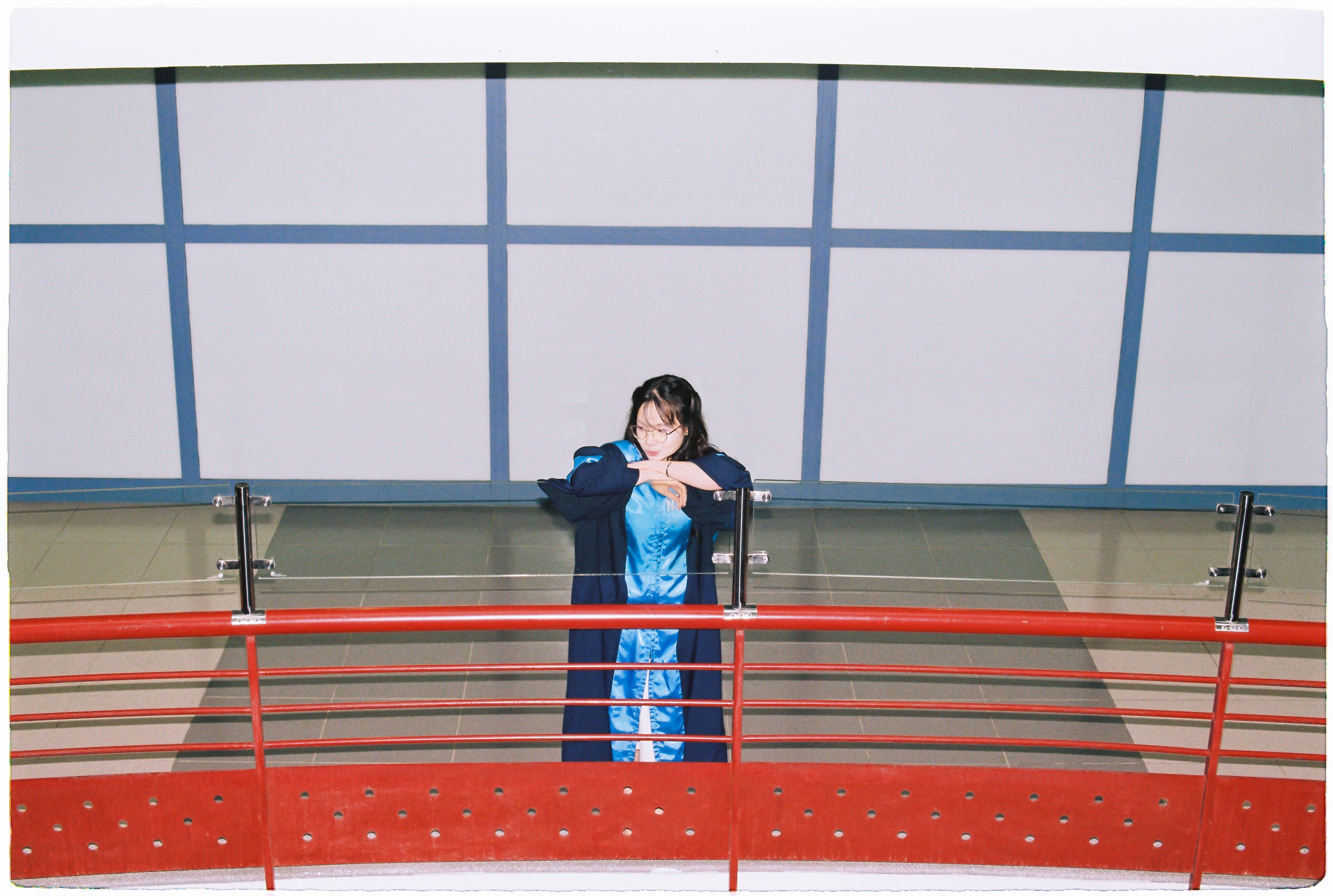 Young woman in blue gown leaning on red railing indoors, with gray wall background.