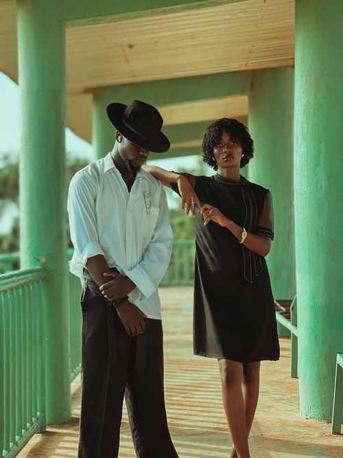 Free Elegant couple in fashion poses on a sunlit balcony with green pillars and vintage attire. Stock Photo