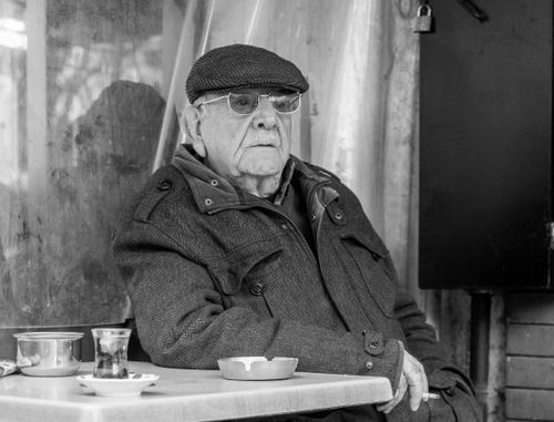 Free Black and white portrait of a senior man in a flat cap, seated outside a café with empty tea glasses on the table. Stock Photo