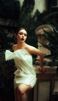 Stylish woman posing gracefully in a white dress surrounded by lush indoor plants, exuding elegance.