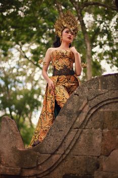 Woman in intricate golden traditional attire posing outdoors with trees in the background.