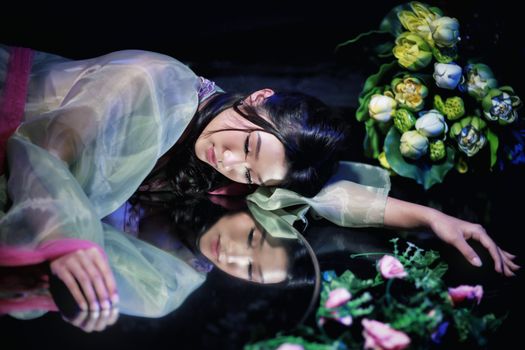 A serene woman in traditional dress lies peacefully by her reflection with vibrant flowers.