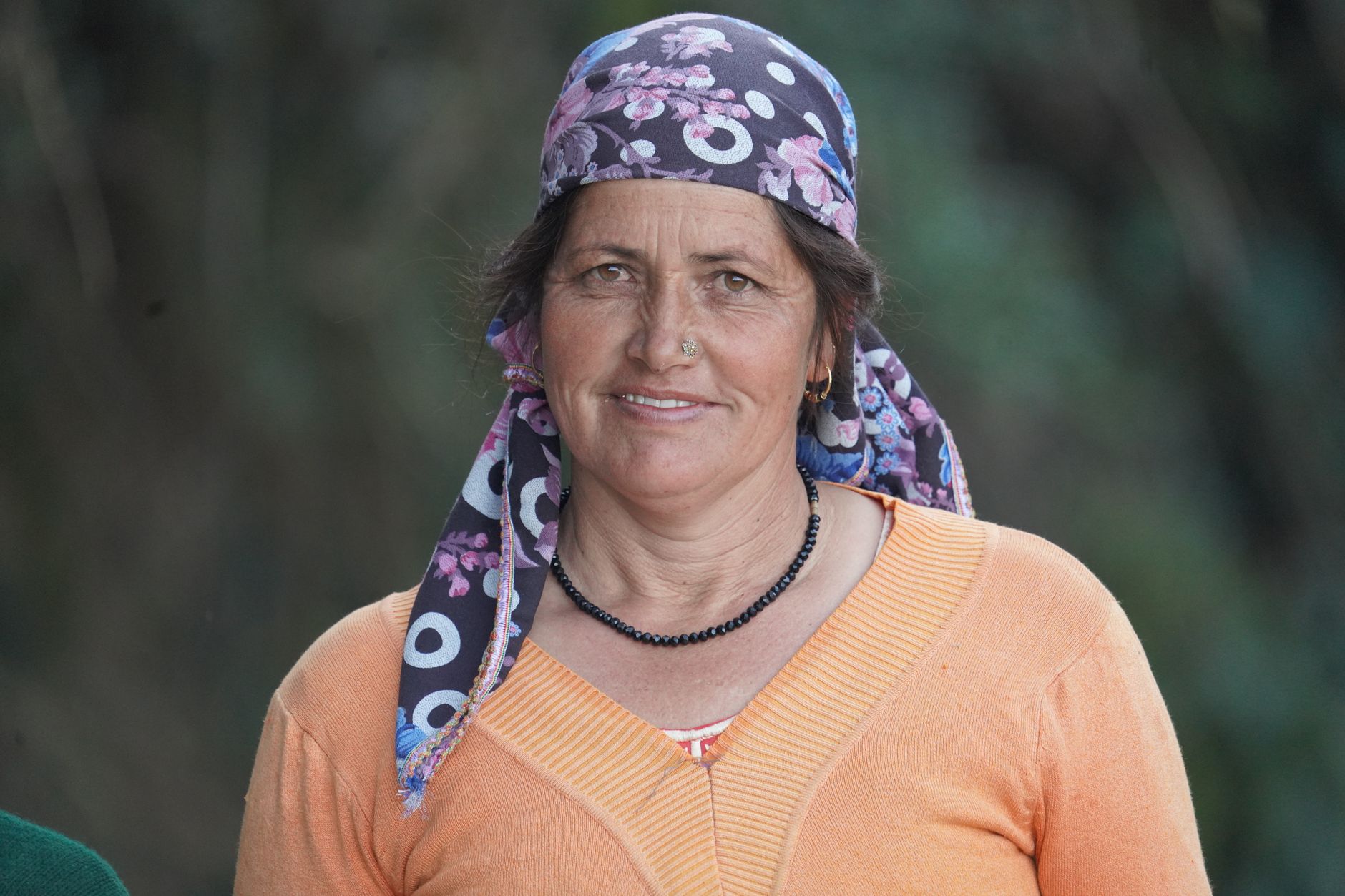 A middle-aged woman with a floral headscarf smiles warmly outdoors.