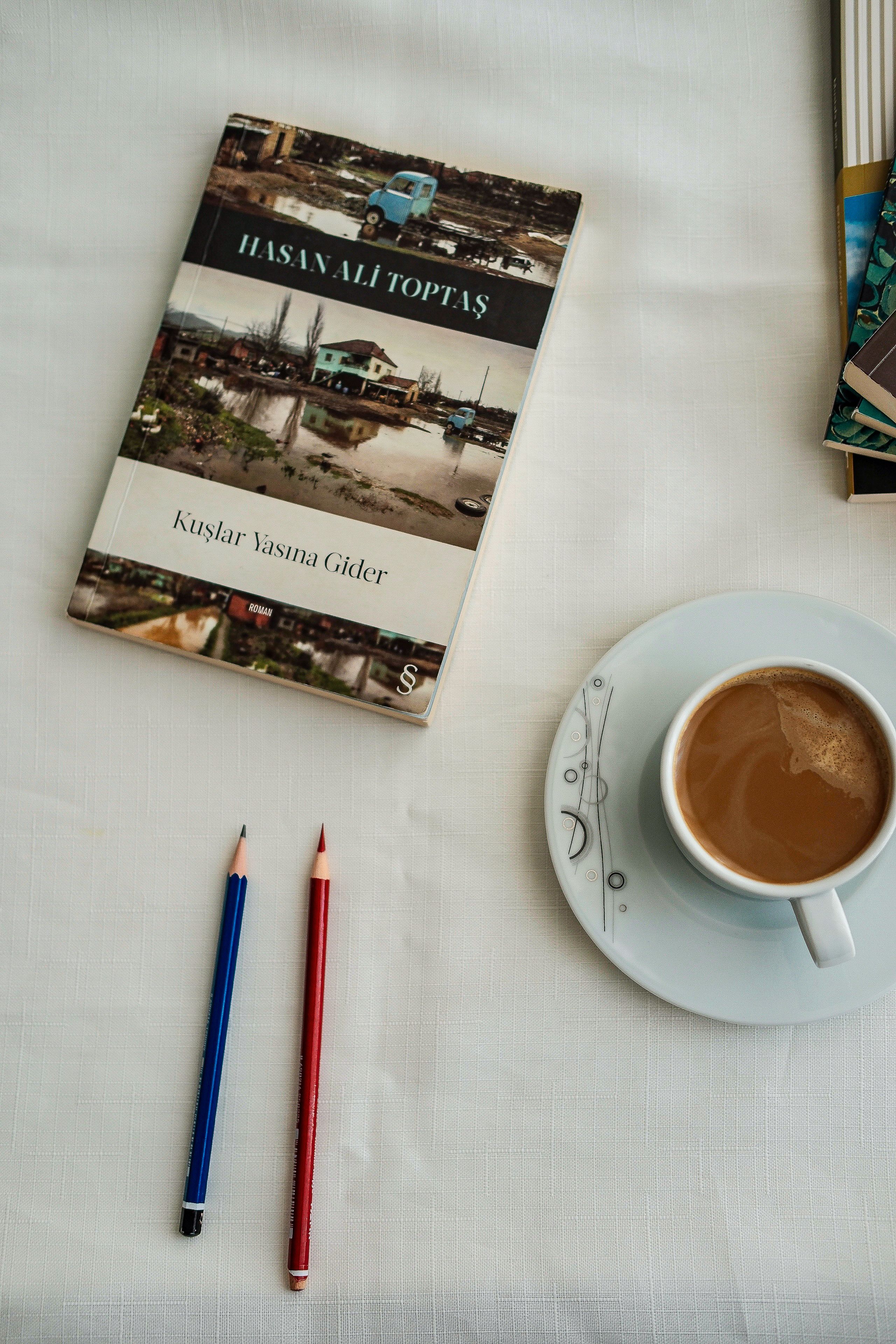 A book, coffee cup, and colored pencils on a white tablecloth create a cozy reading scene.