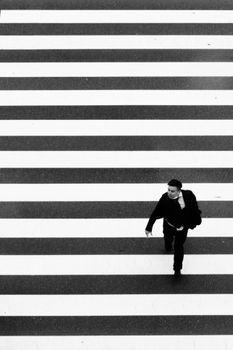 A lone man crossing a zebra crossing in a black and white urban setting.