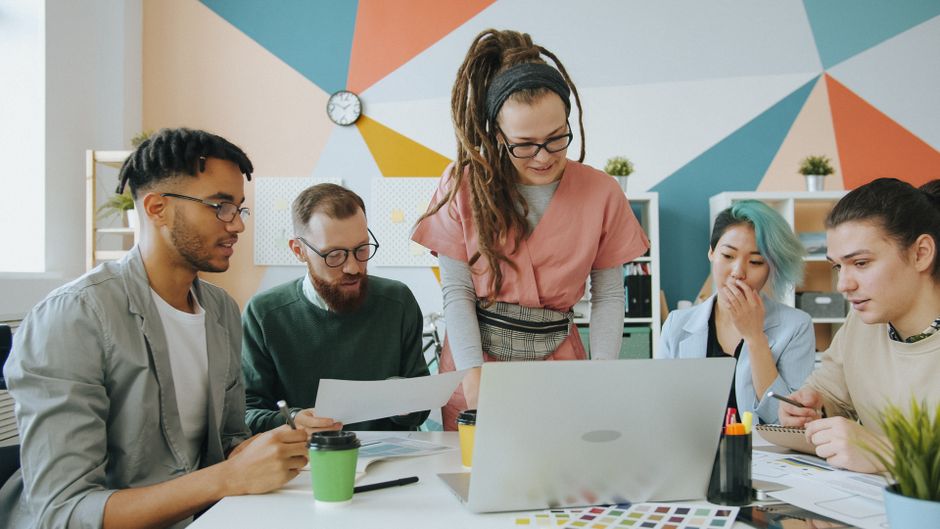 Un equipo diverso de jóvenes adultos colabora alrededor de un ordenador portátil en un entorno de oficina moderno y colorido.
