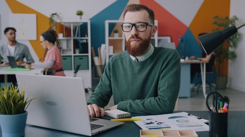 Free Man with glasses and beard working on laptop in colorful office. Stock Photo