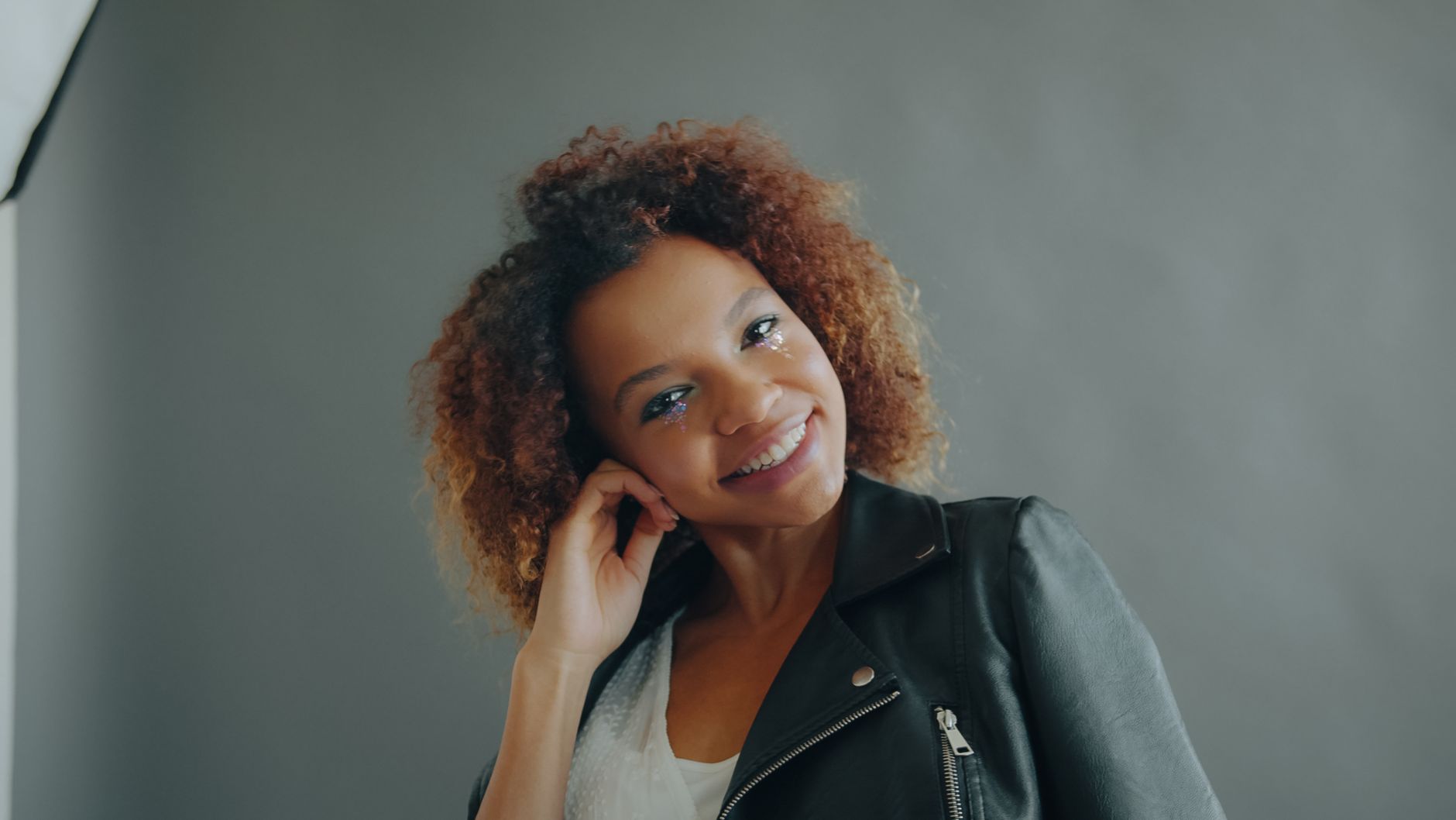 A cheerful woman with curly hair poses in a leather jacket against a gray background.