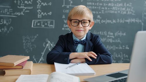 Free Young boy in suit and glasses sitting at school desk with chalkboard behind. Stock Photo