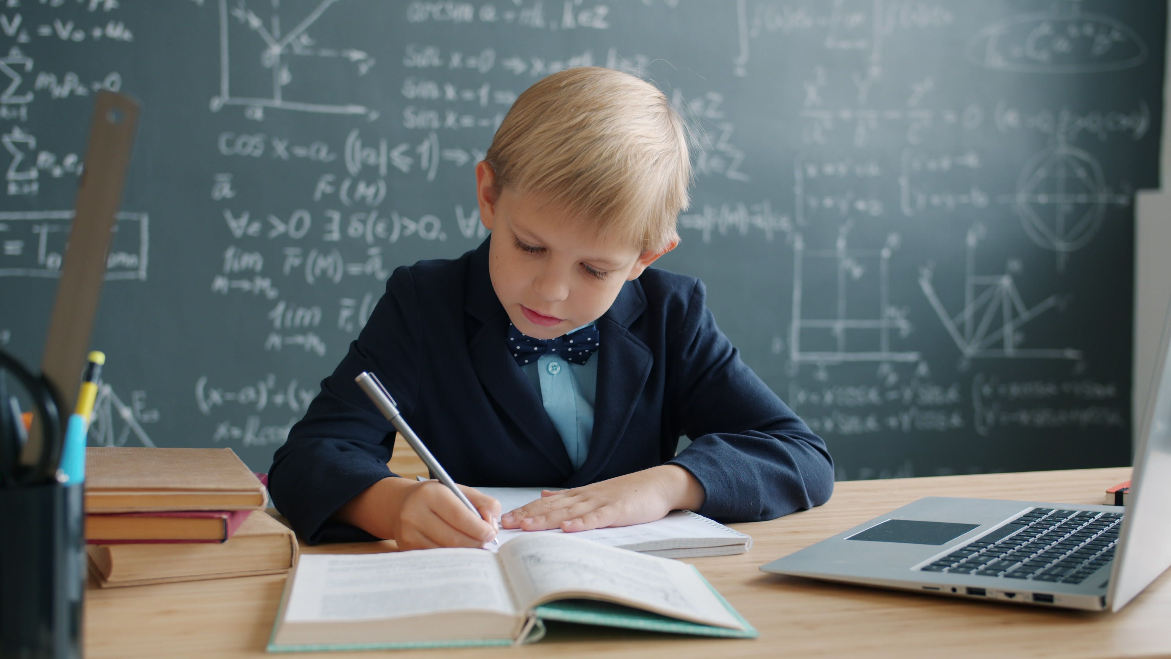A young student diligently works on an assignment in a classroom with a laptop nearby.