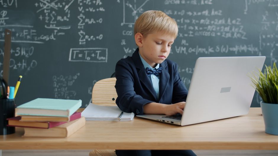 Young boy in suit uses laptop in math classroom with chalkboard.