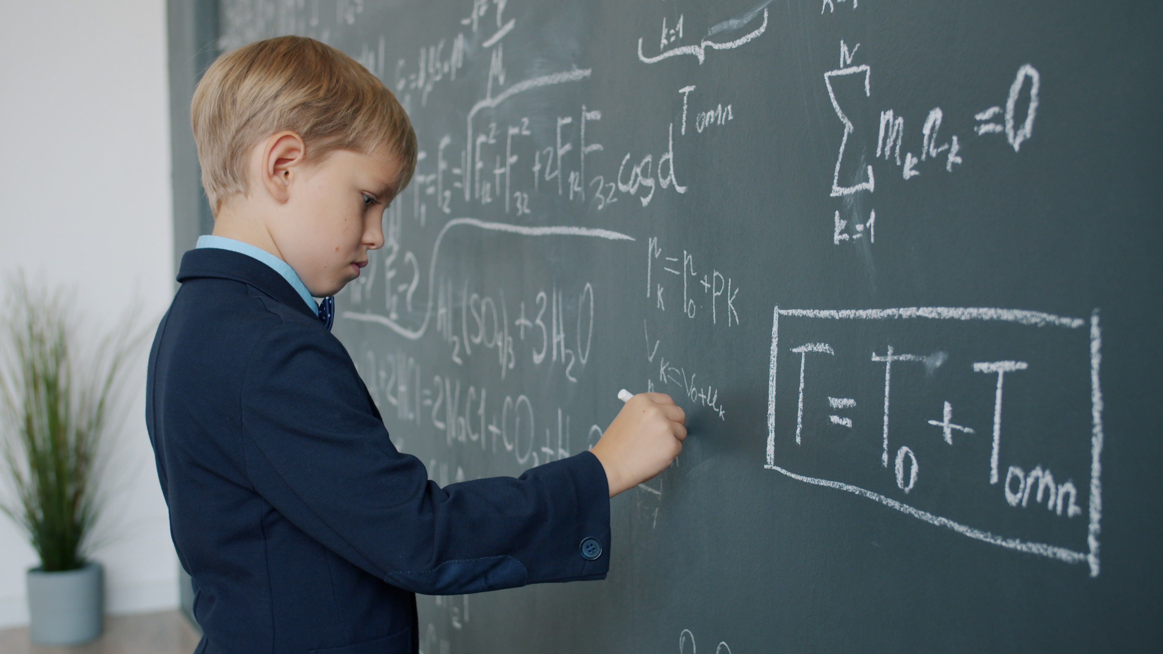 A boy in a suit writing complex math equations on a classroom blackboard.