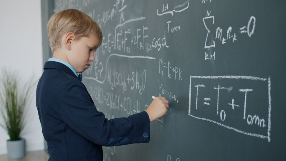 A boy in a suit writing complex math equations on a classroom blackboard.