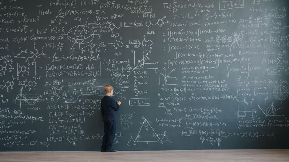 Child in suit writing complex equations on a large blackboard.