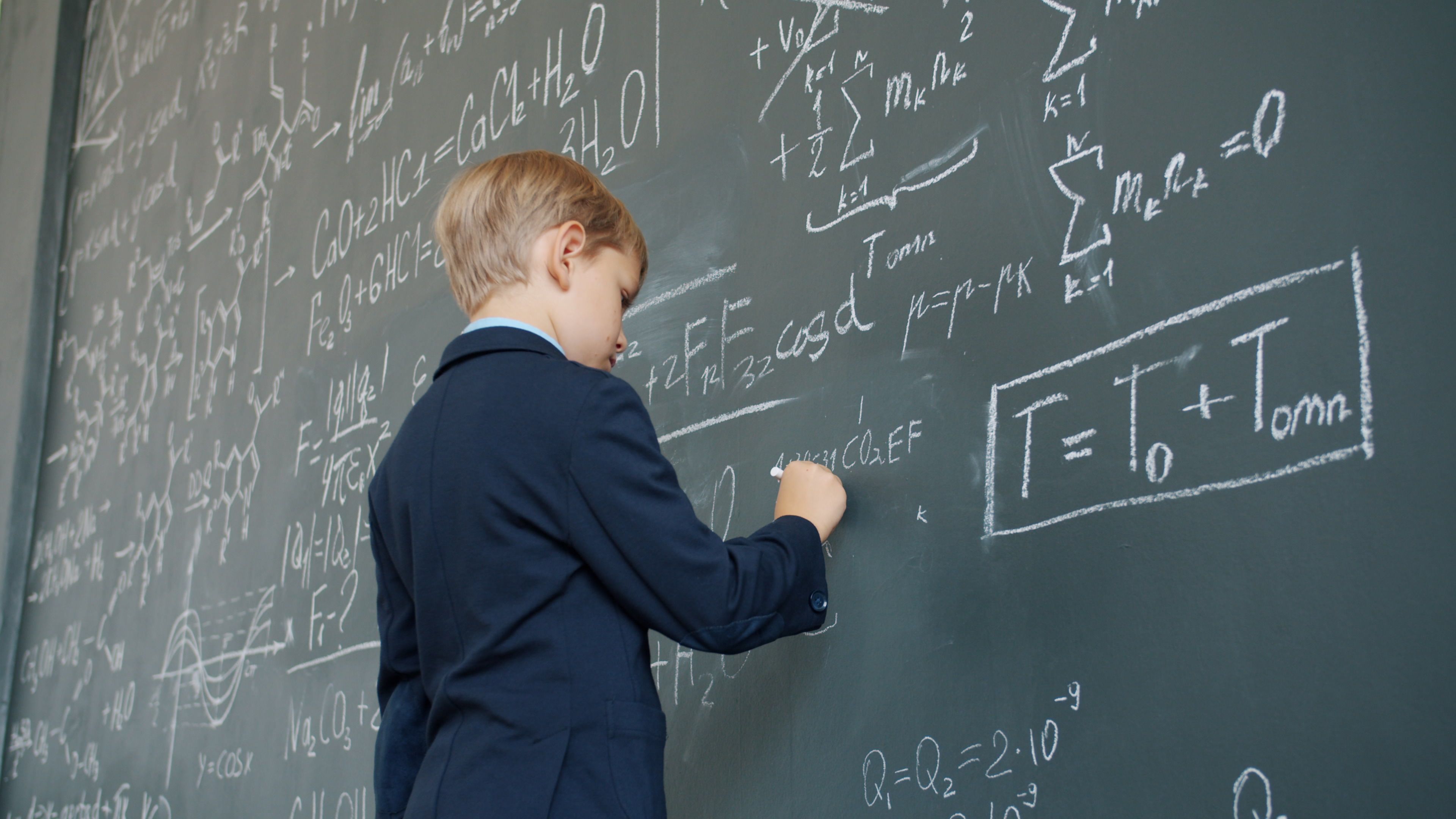 Boy in suit writing math equations on classroom chalkboard.
