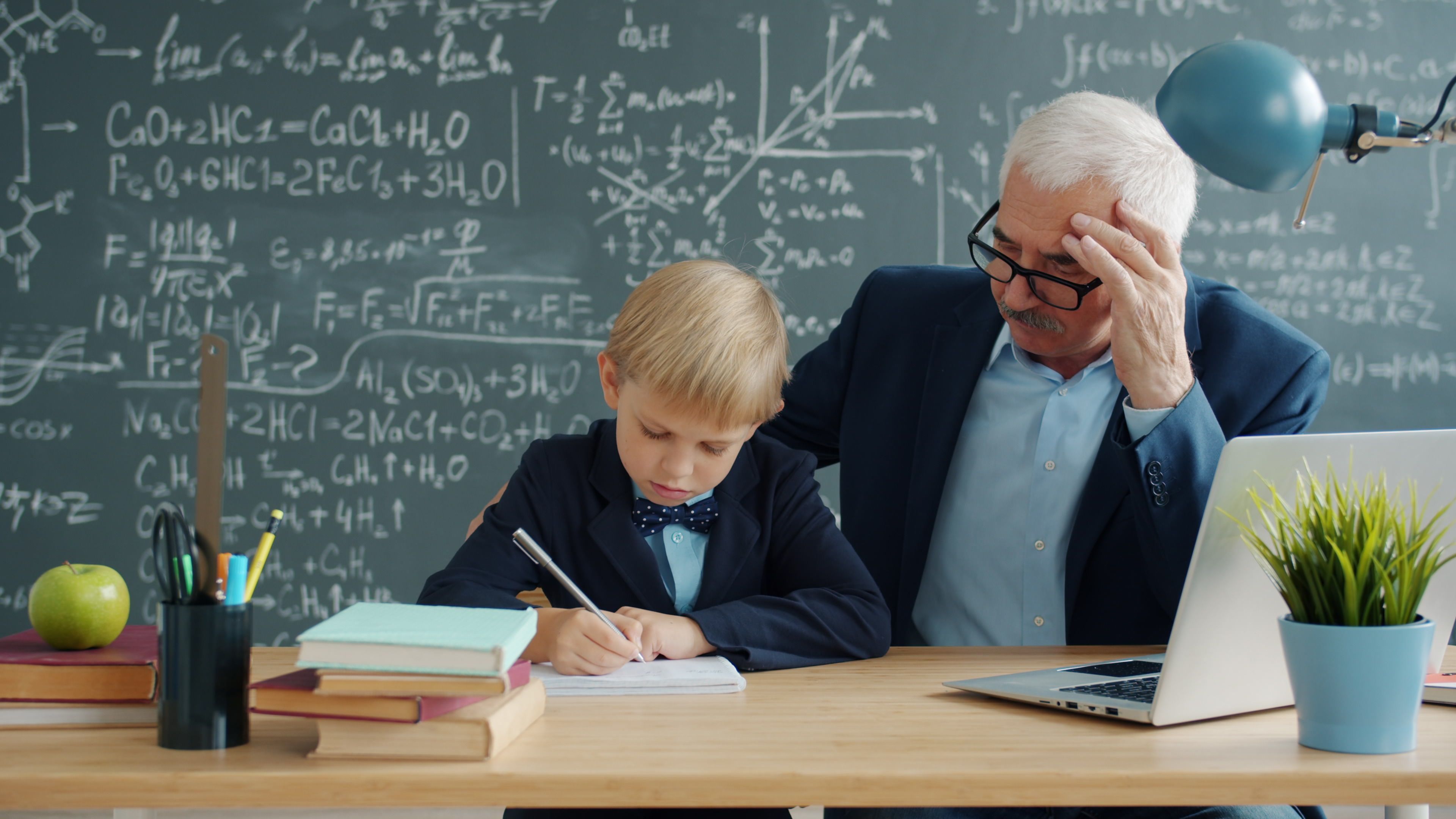 Elderly teacher helping a young student with homework in a classroom setting.