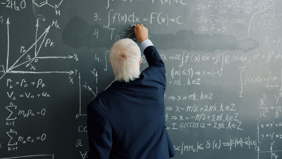 An elderly professor writing complex math equations on a classroom chalkboard.
