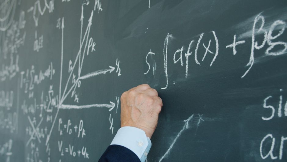 Close-up of a hand writing complex mathematical equations on a chalkboard.
