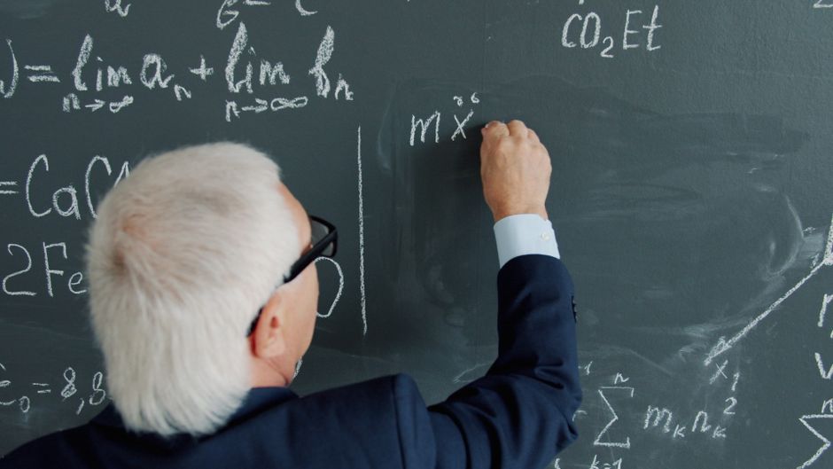 Senior male professor writing complex equations on a chalkboard.