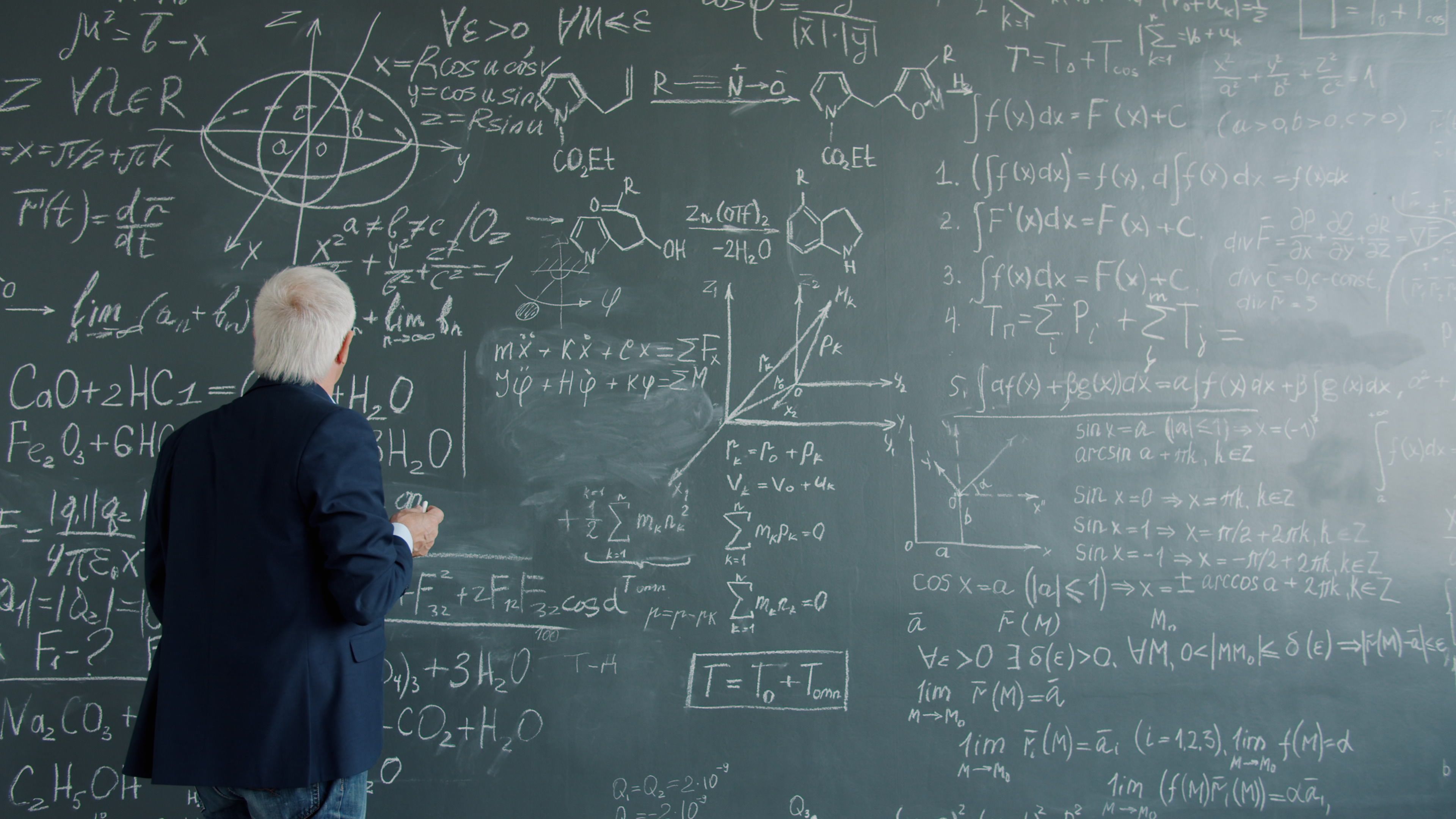 A mature professor writes complex mathematical equations on a large chalkboard.