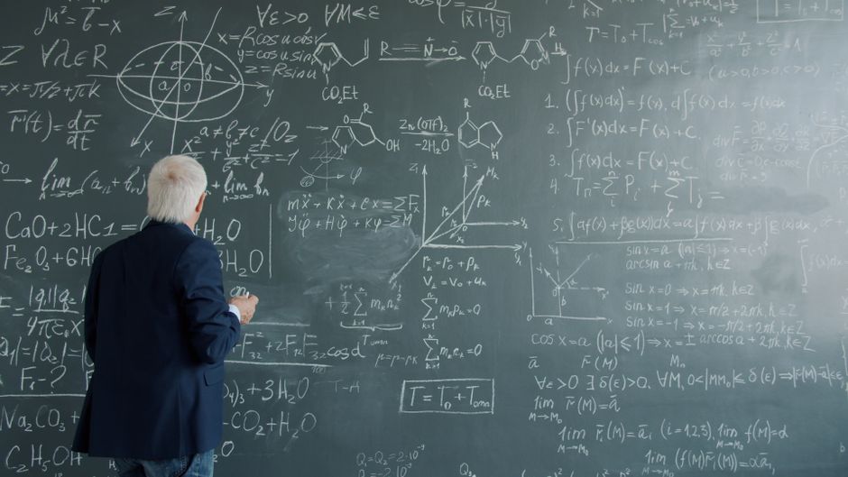 A mature professor writes complex mathematical equations on a large chalkboard.