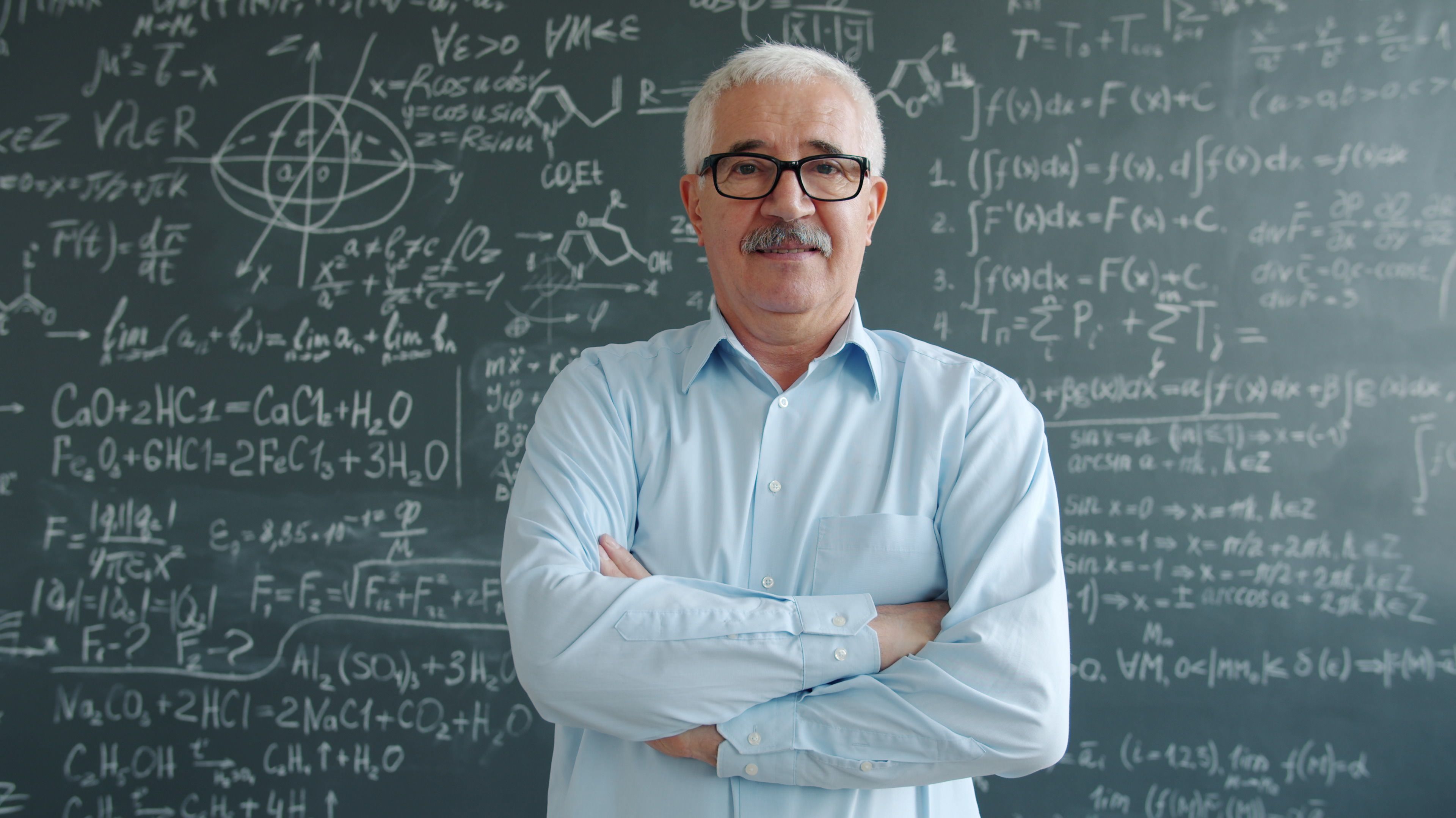 Senior professor with folded arms in front of chalkboard filled with scientific equations, essence of education.