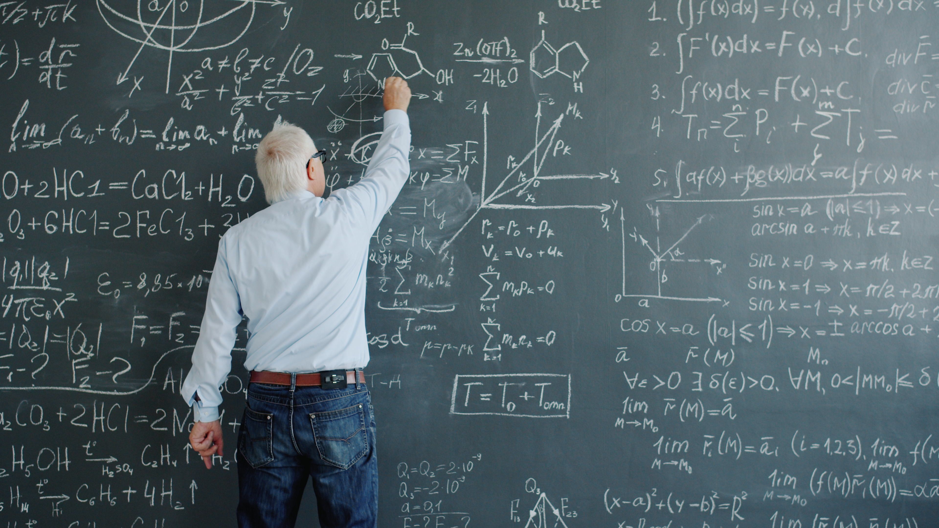 A senior professor writing complex mathematical equations on a blackboard in a classroom.