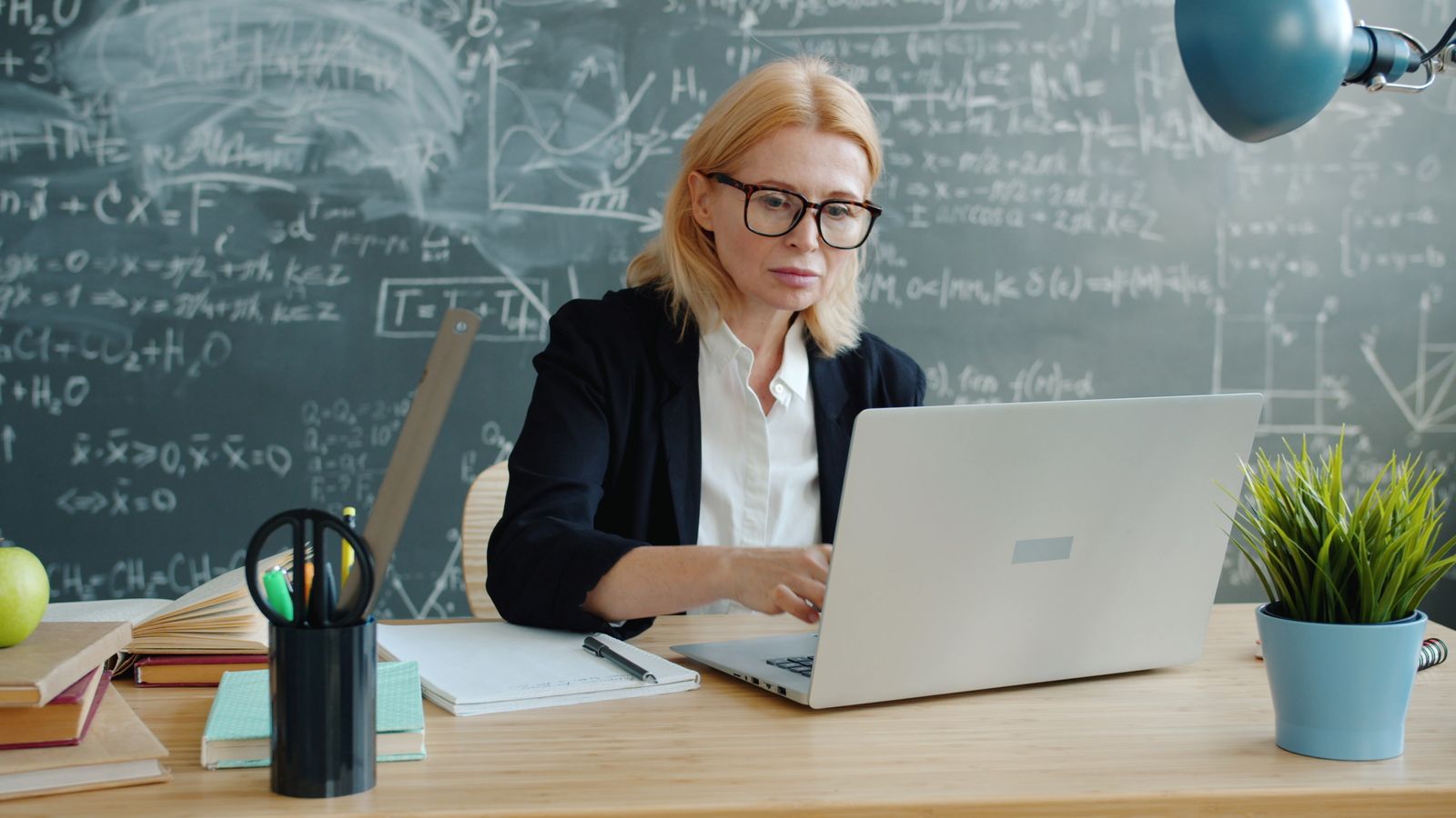 Docente a trabalhar num computador portátil diante de um quadro de sala de aula