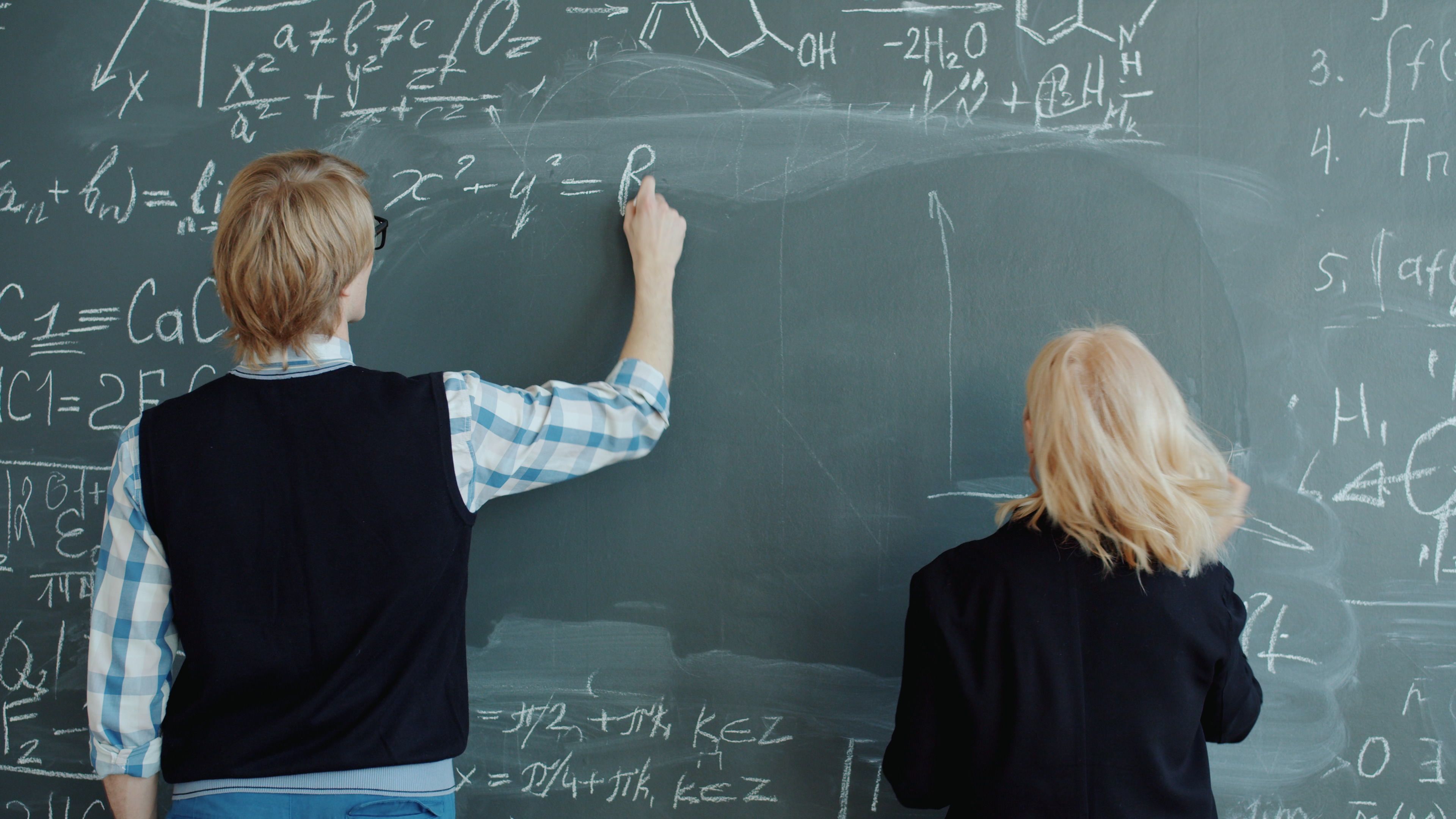 Two students write complex mathematical equations on a large blackboard.