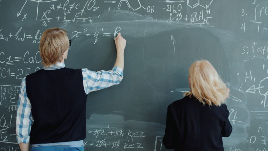 Two students write complex mathematical equations on a large blackboard.