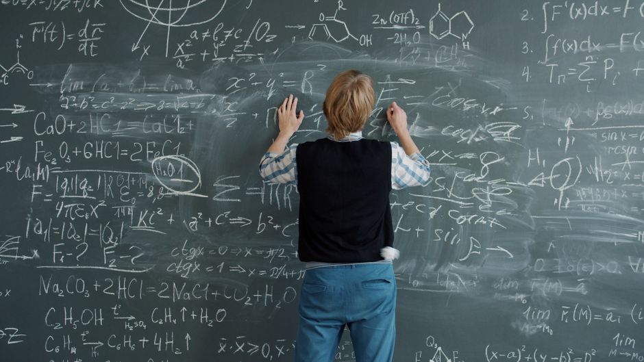 Rear view of teacher solving complex equations on chalkboard in a classroom.
