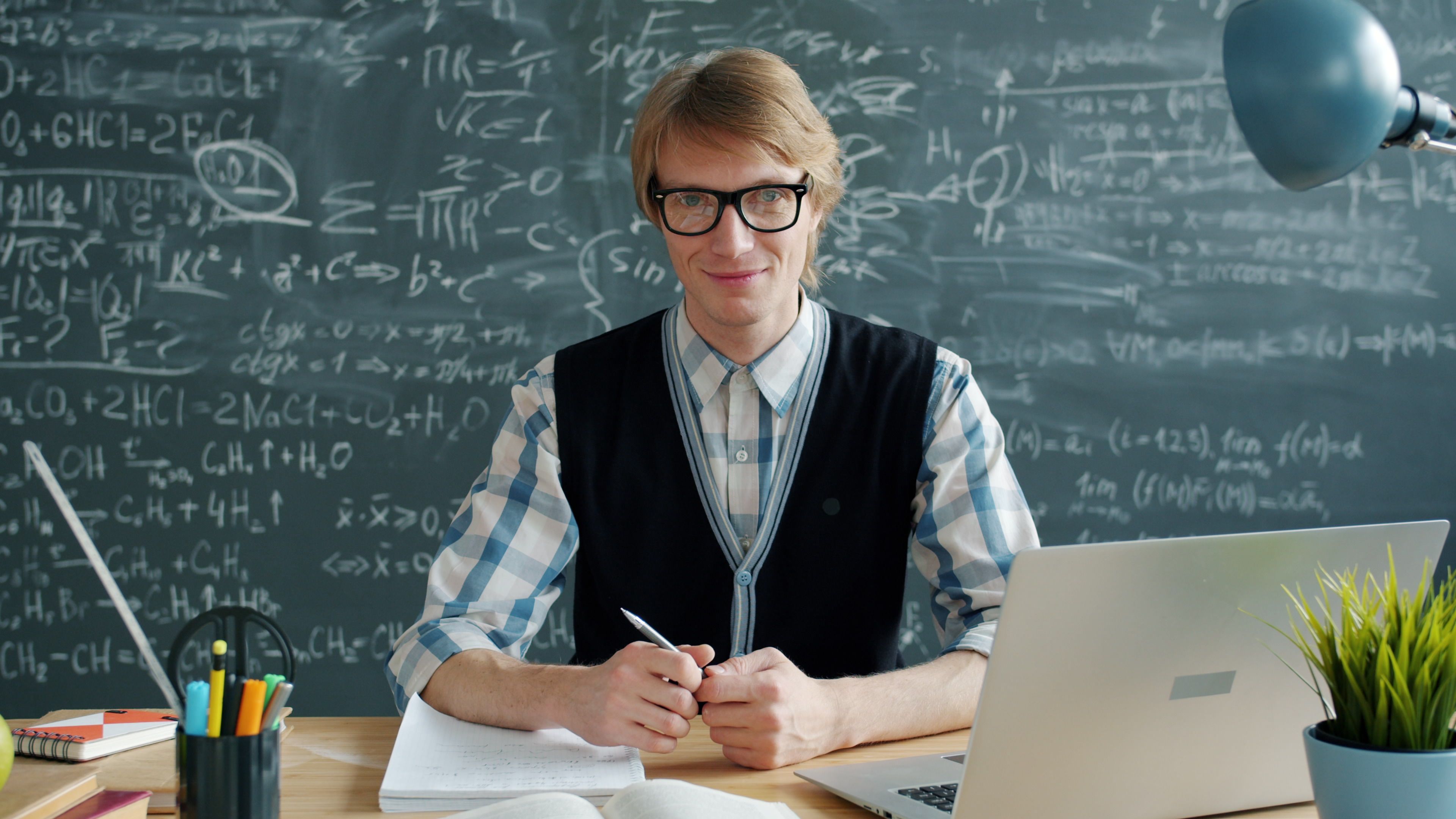 Educator in plaid shirt sitting at desk in front of chalkboard filled with equations