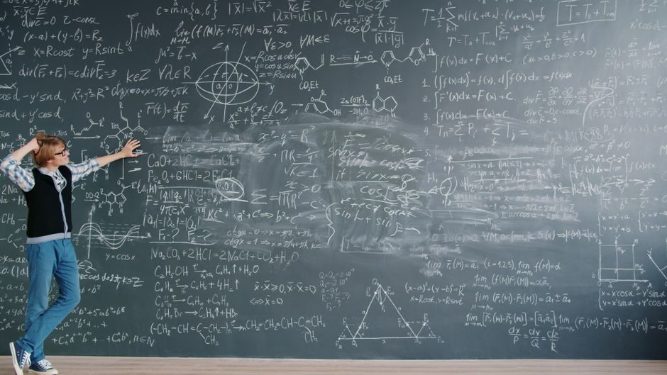 Student stands by chalkboard filled with complex math equations in a classroom setting.