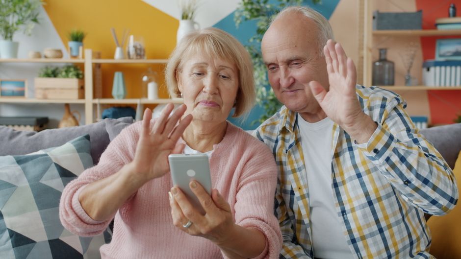 高齢の夫婦がスマートフォンを使ってビデオチャットをしながら、手を振って挨拶している。