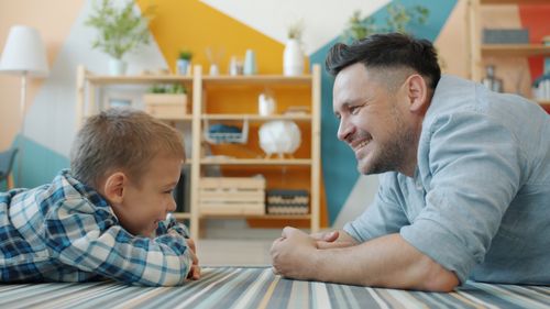 Free Father and son enjoying a playful moment indoors, lying on the floor and smiling at each other. Stock Photo