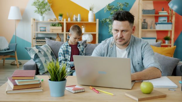 Father on laptop working from home, son playing in colorful living room.