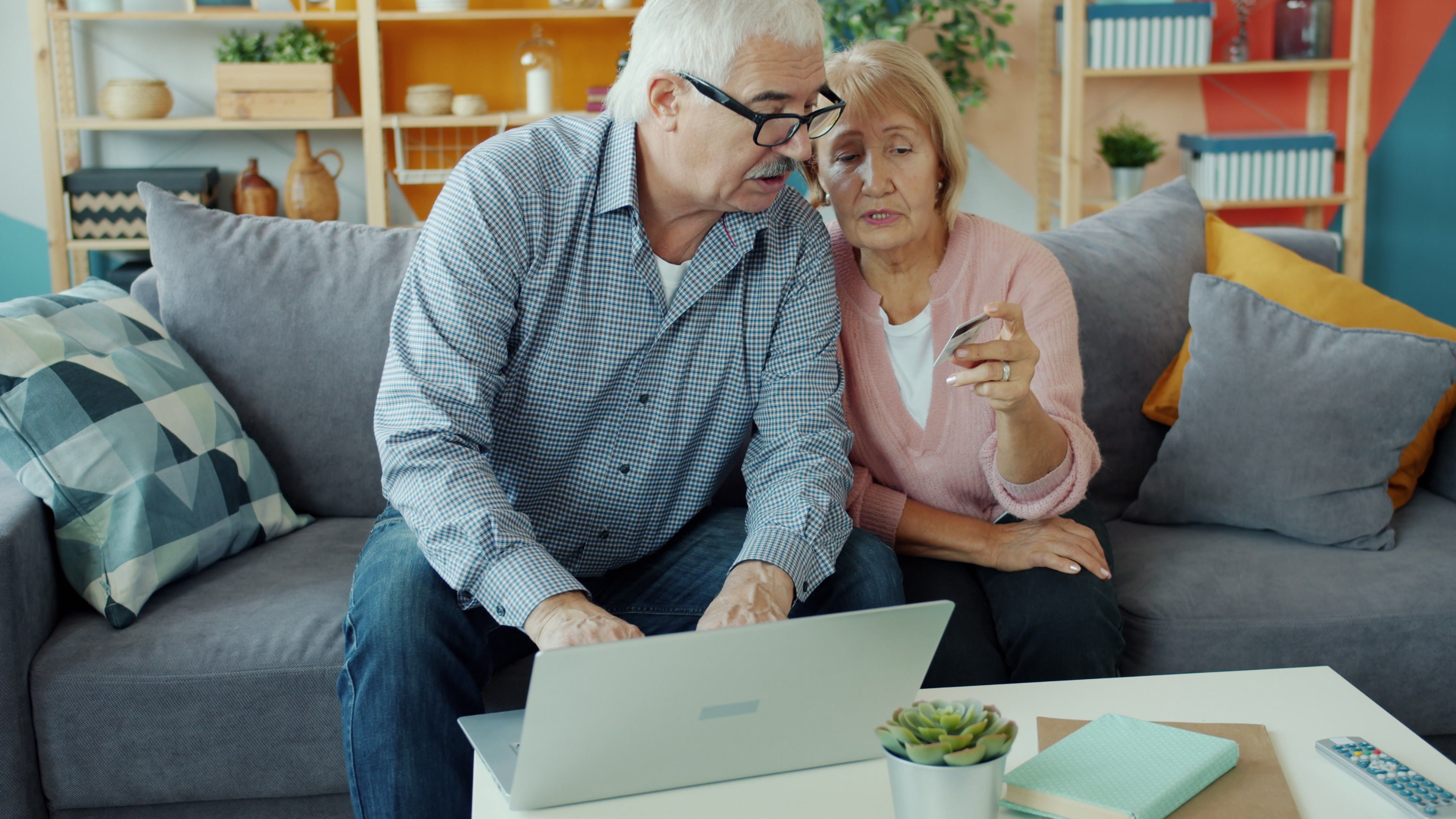 Elderly couple using a laptop and credit card in a cozy living room.