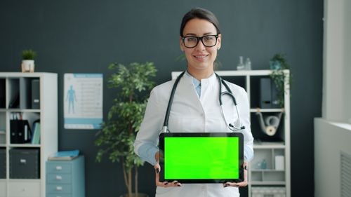 Free Female doctor smiling and holding a tablet with green screen in a modern office. Stock Photo