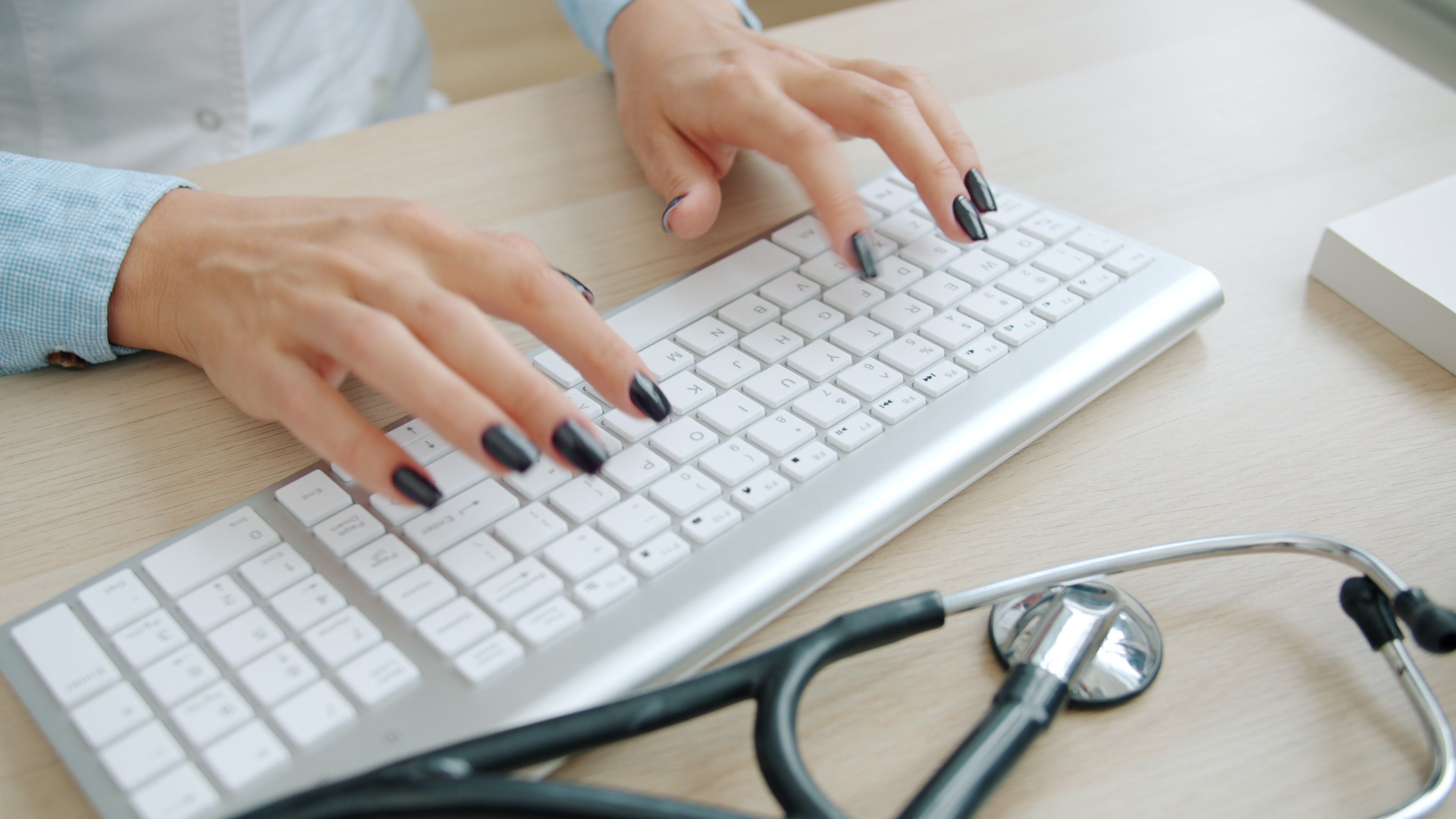Close-up of doctor typing on computer with stethoscope on desk.