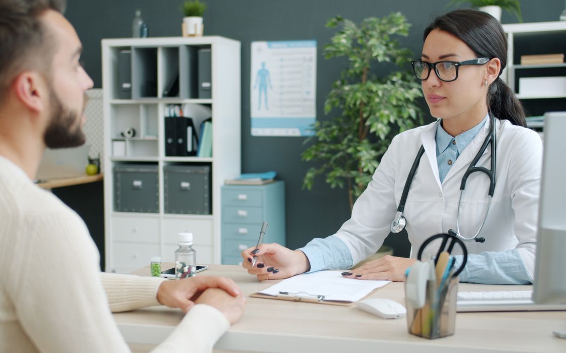 Doctor in a white coat discussing treatment with a male patient across a desk in a modern clinic office
