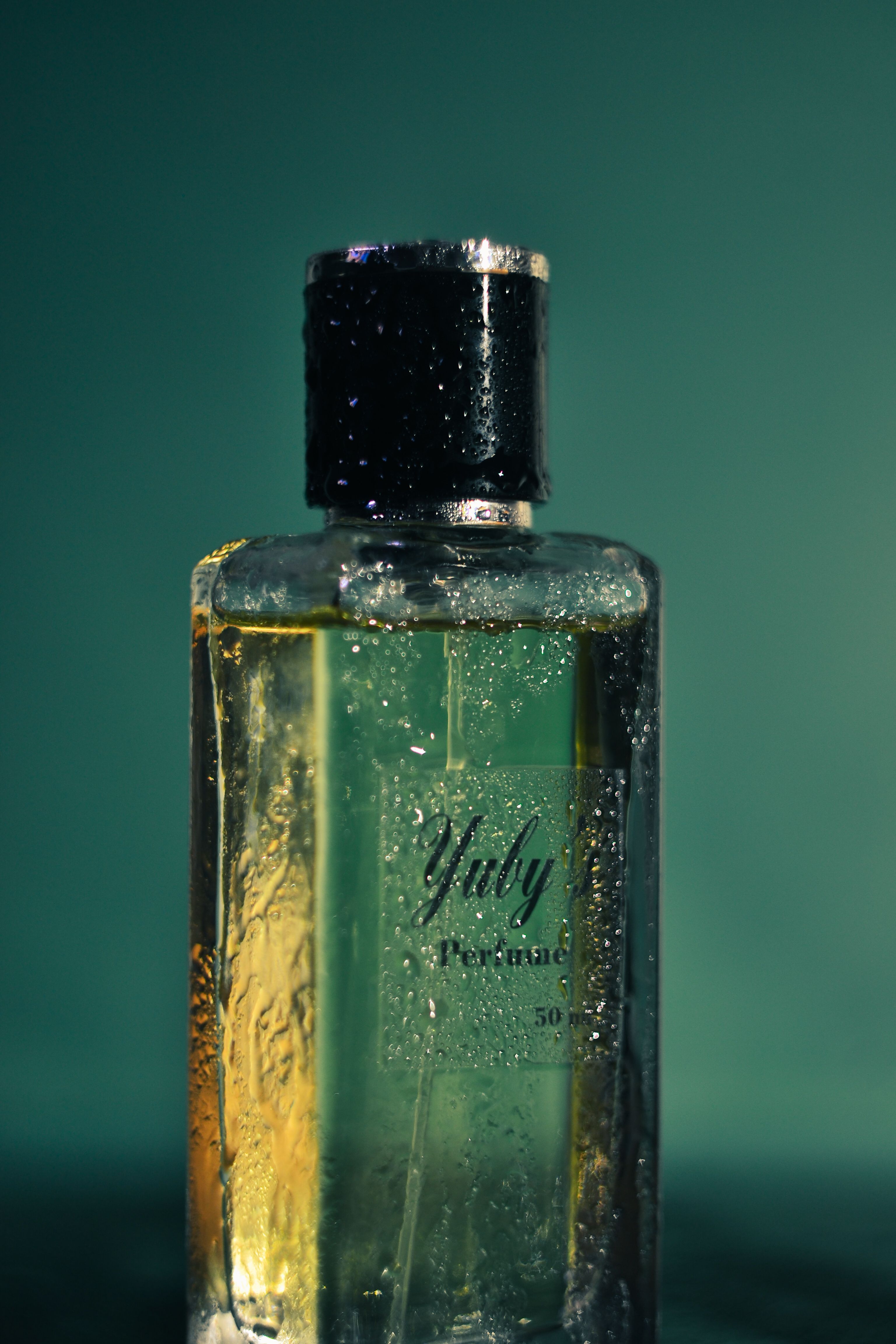 Close-up of a wet perfume bottle with 'Yuly' label, capturing shimmering light on green backdrop.