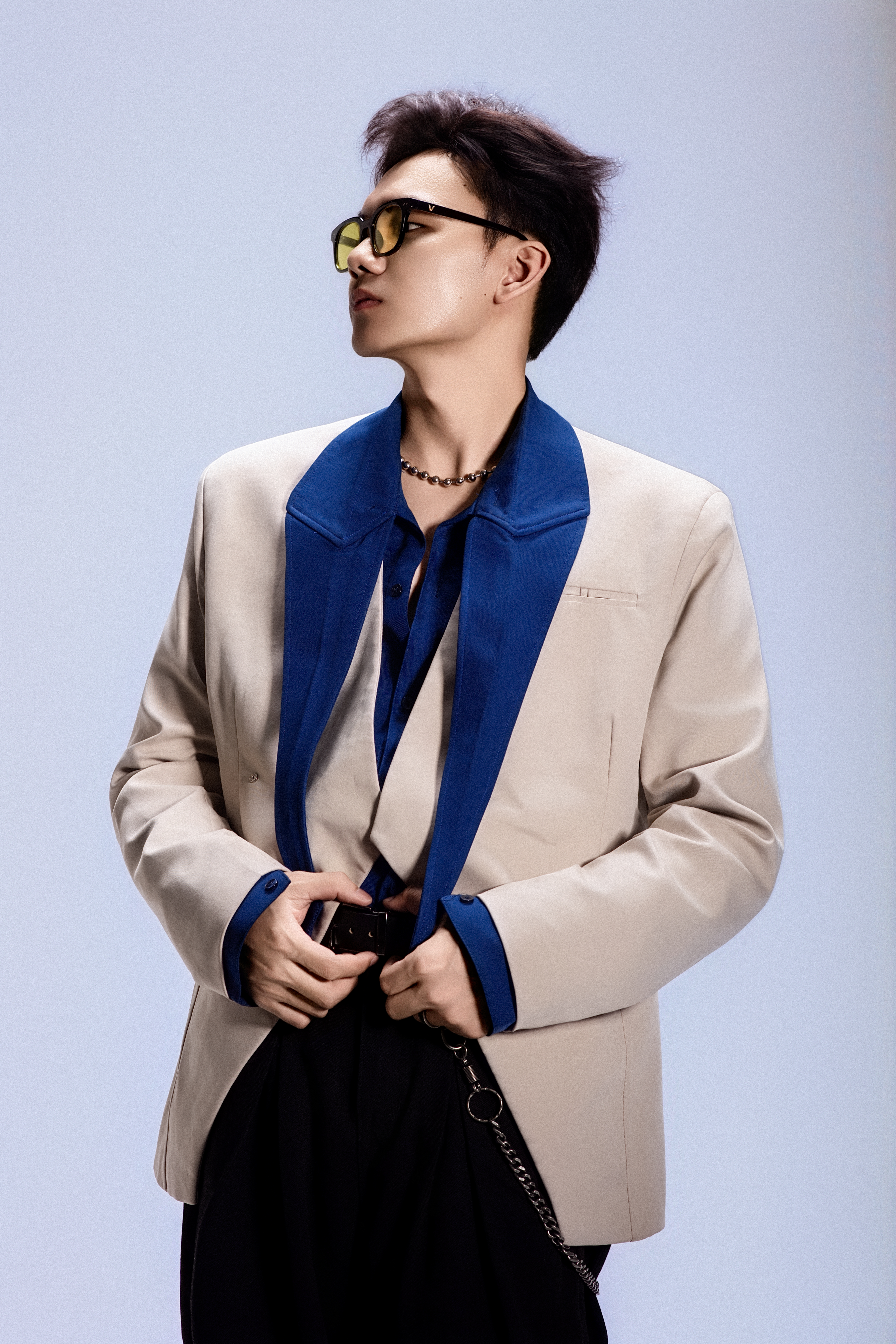 Fashion-forward young man in sunglasses and beige jacket posing coolly in studio