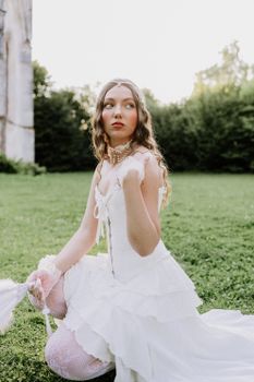 A young woman in a vintage white gown sits on grass during the day.