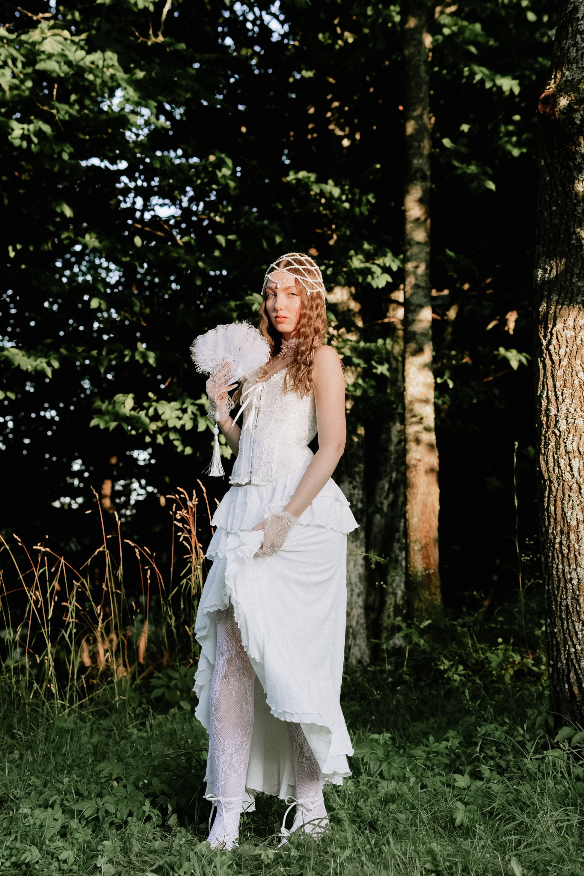 Elegant woman in a vintage white dress stands outdoors with a fan, surrounded by nature.